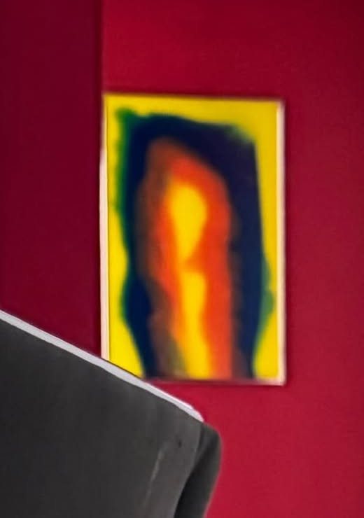 Espelho retangular em amarelo, azul, laranja e vermelho numa parede vermelha, com o encosto de uma cadeira em primeiro plano.